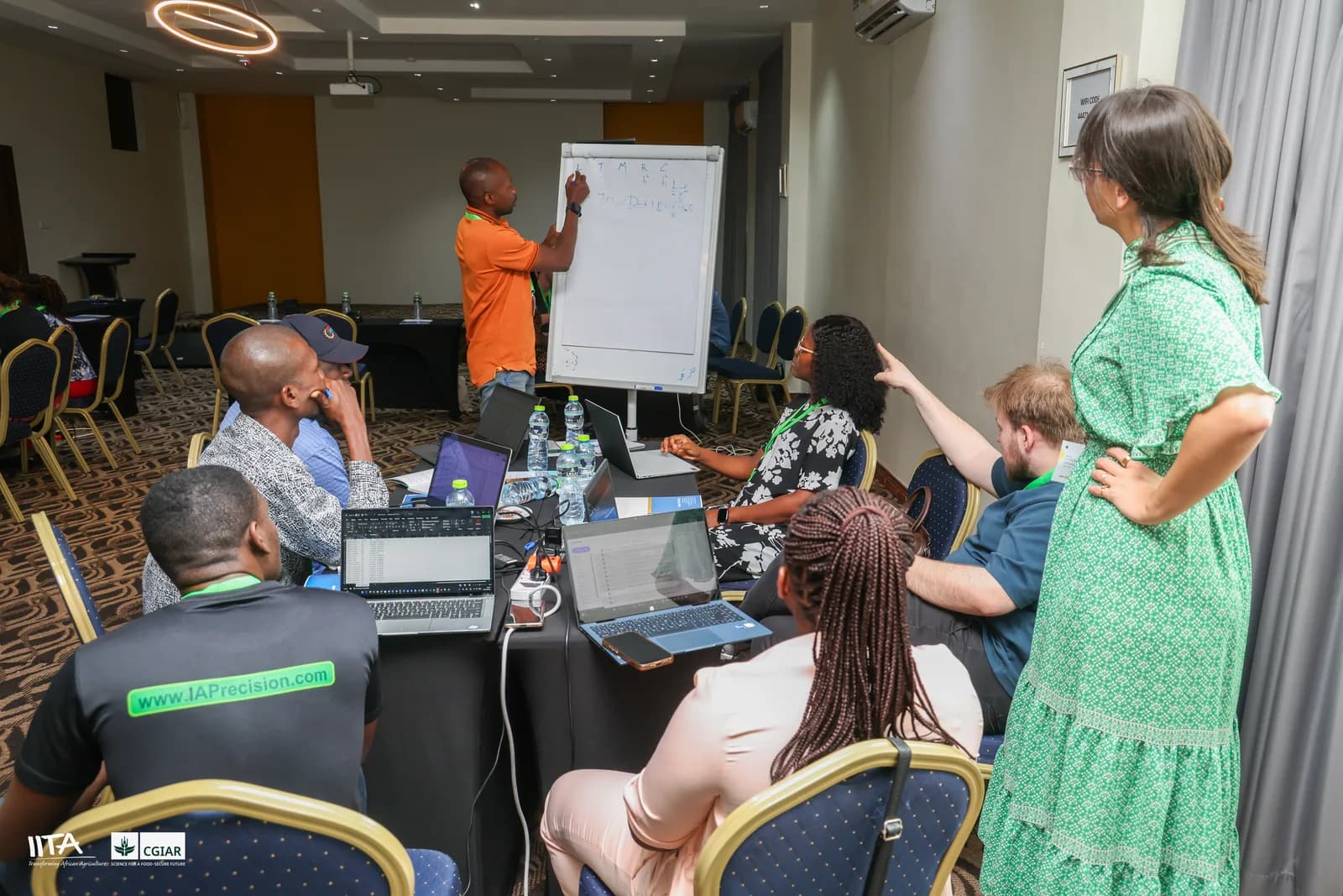 A trainer at a whiteboard working through an analysis question with a small group of participants gathered around their laptops